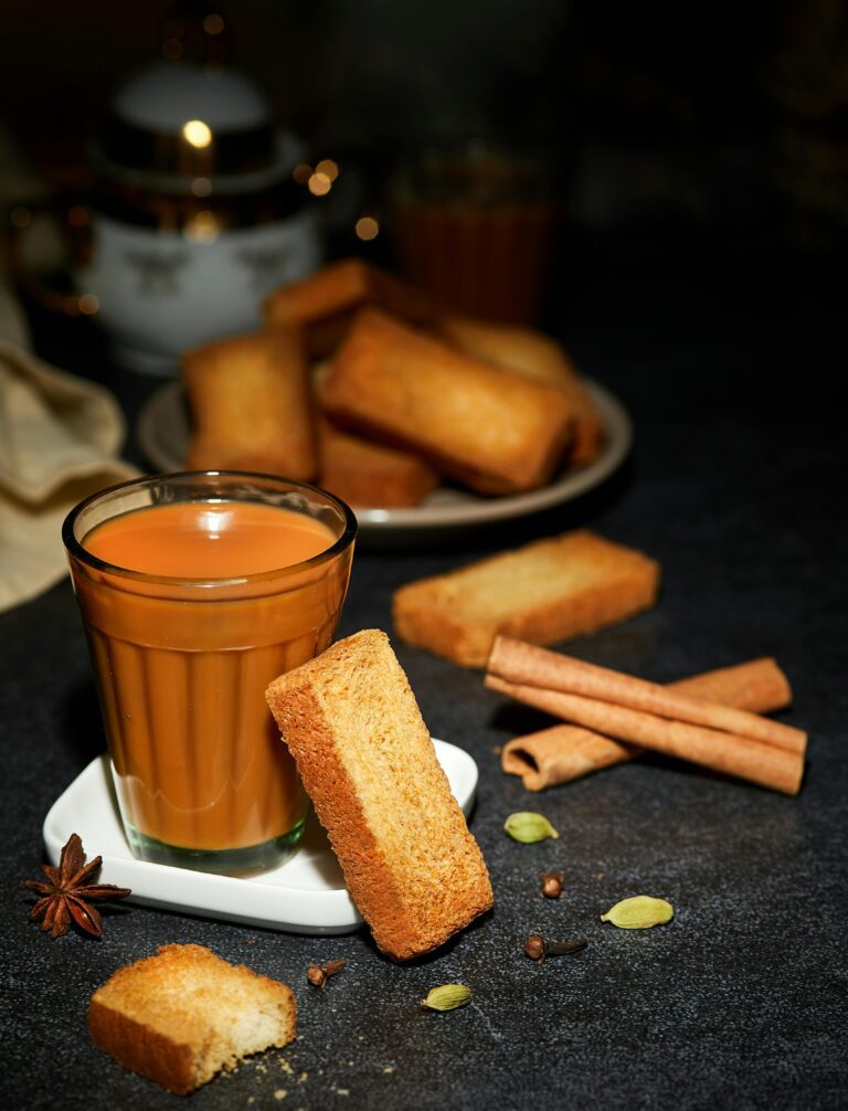 A cup of cardamom chai tea accompanied by biscotti, with spices like cinnamon sticks and star anise scattered around on a dark surface