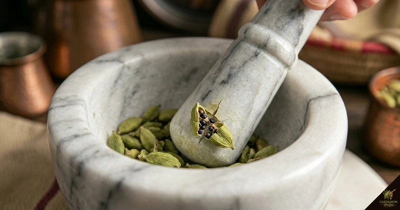 Bruising green cardamom pods with mortar and pestle — correct doodh elaichi technique showing cracked pods with seeds visible