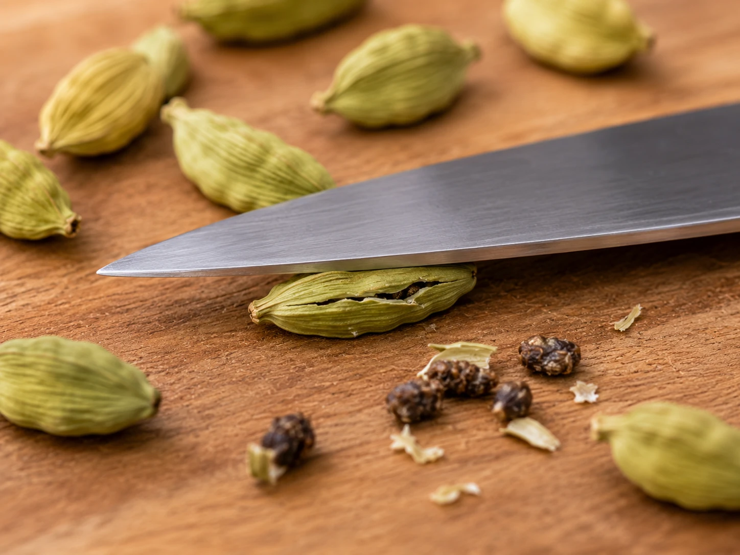 Cardamom pod cracked open with the flat of a knife on a wooden board revealing the seeds