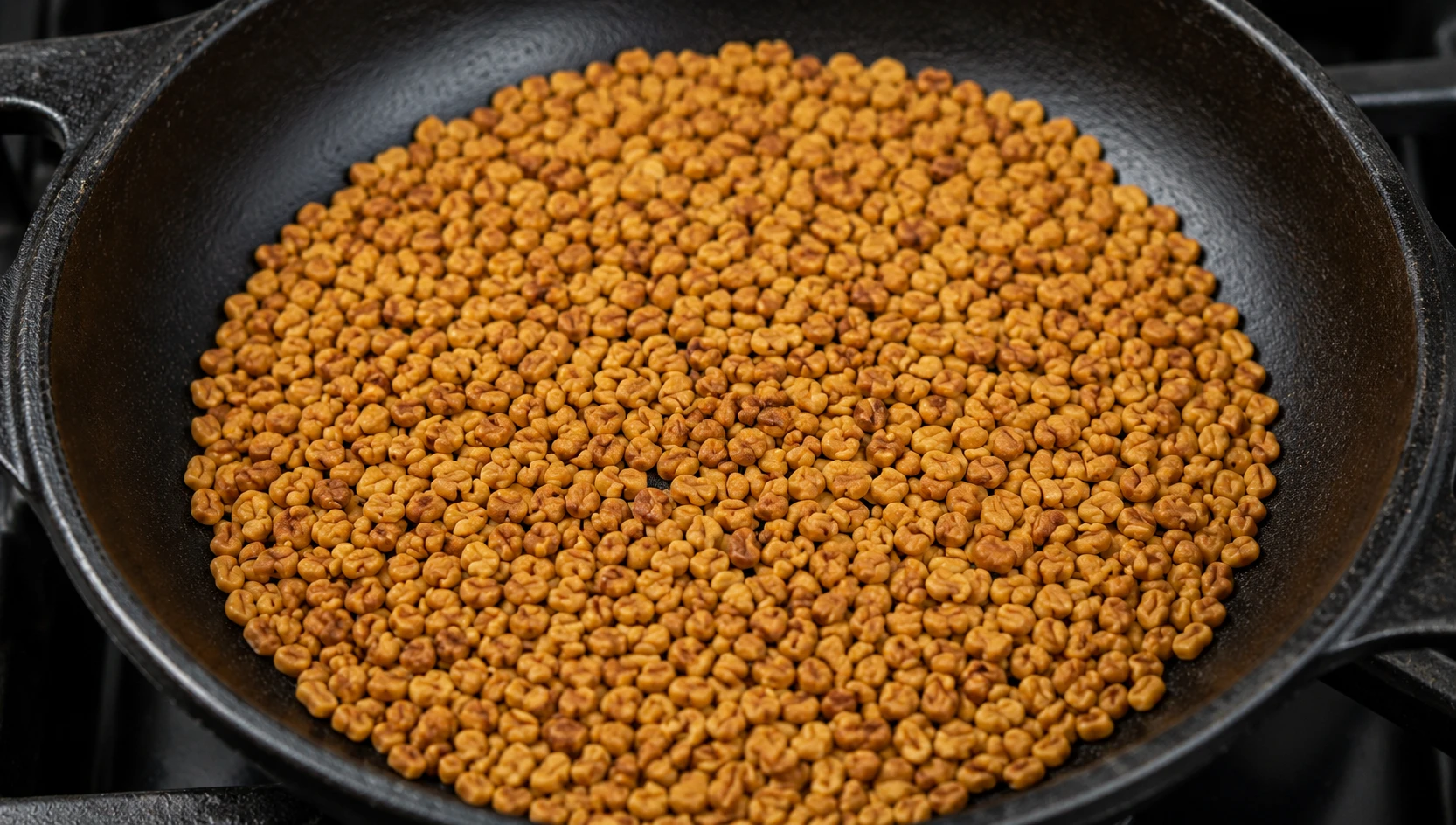 Fenugreek seeds dry-roasting in a cast iron pan, turning golden-brown — the maple-caramel aroma of sotolone is released during roasting