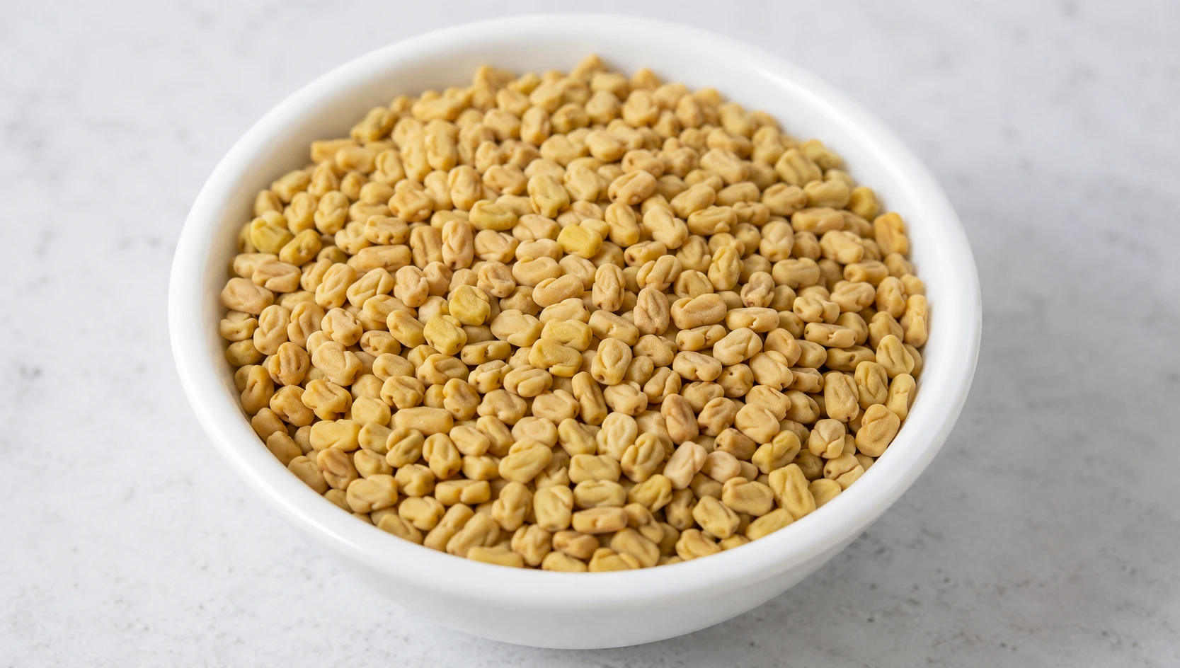 Raw uncooked fenugreek seeds in a small white ceramic bowl before any heat is applied — pale yellow-brown colour